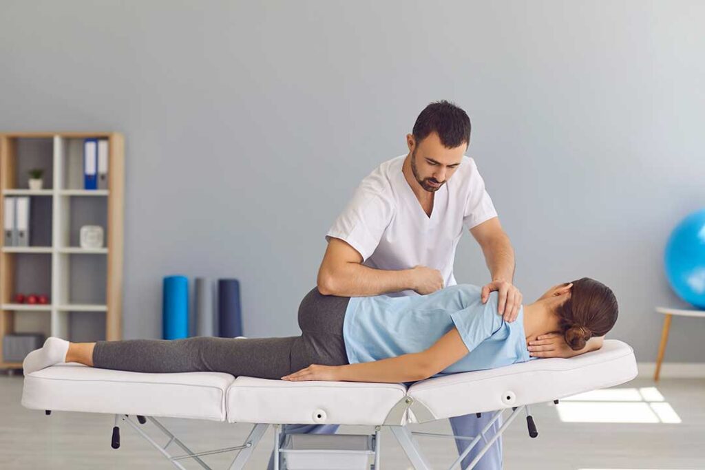 A person gently stretching their knee on a living room mat with a water bottle and light weights nearby.